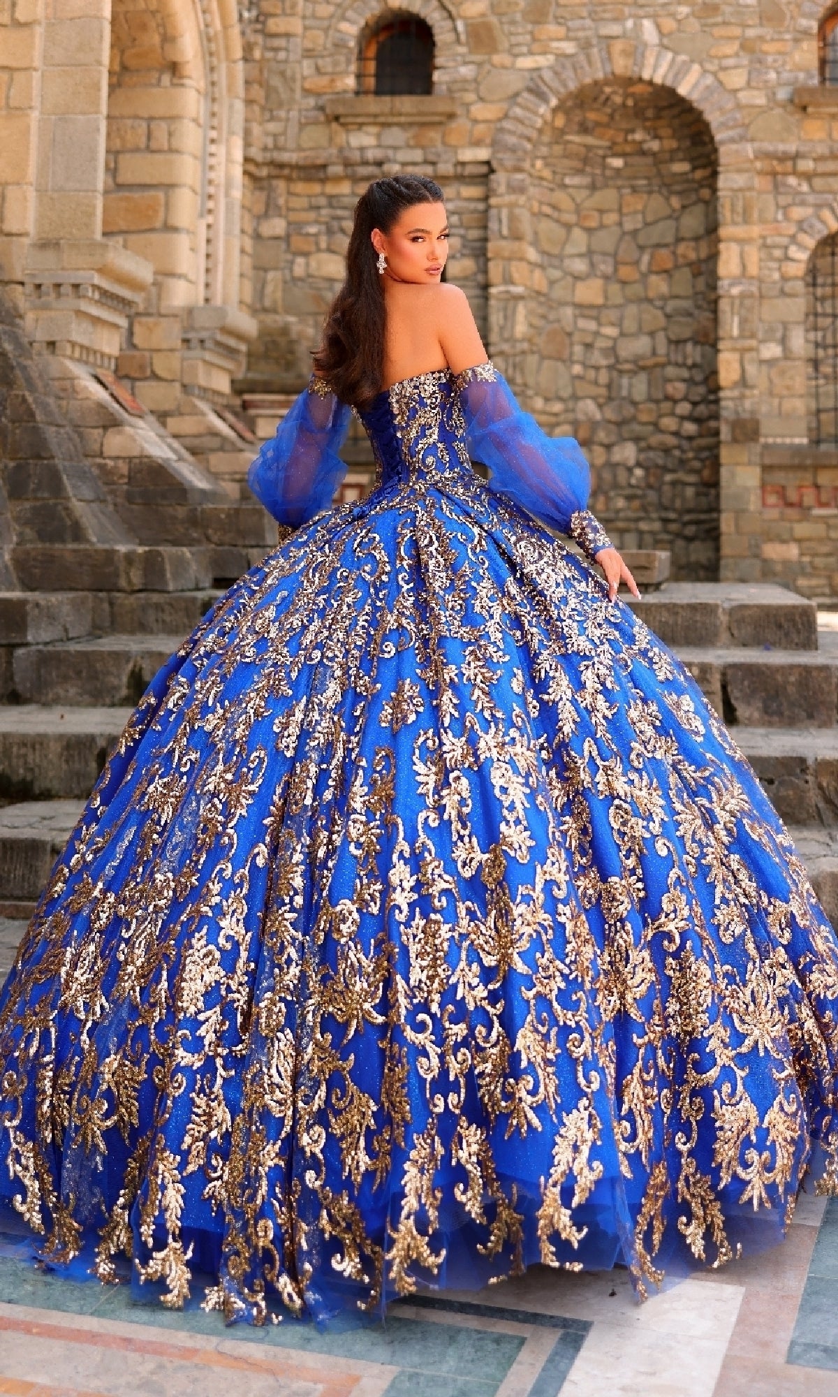 A woman stands on stone steps wearing the Amarra 54238 Sequin Quinceanera Dress, a vibrant blue strapless gown with gold embroidery and a corset bodice, posing before a stone building with arched doorways.